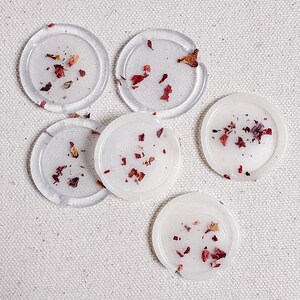 May include: Six clear round coasters with dried rose petals embedded inside. The coasters are arranged in a semi-circle on a white fabric background.
