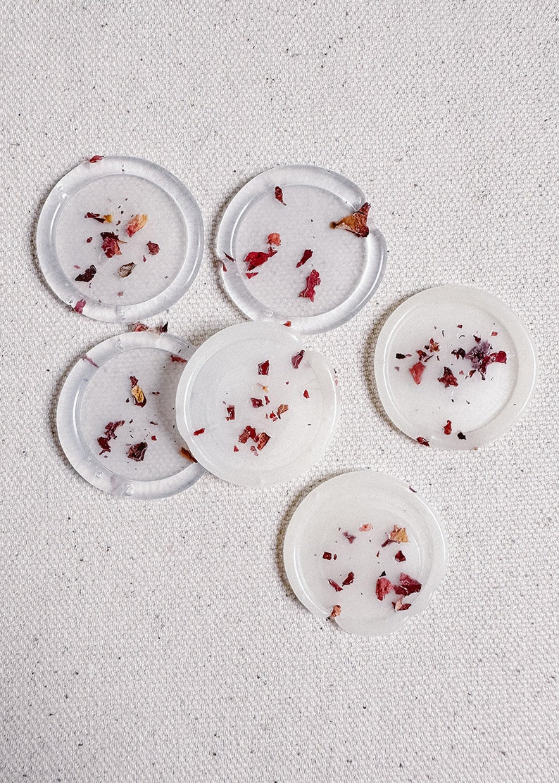 May include: Six clear round coasters with dried rose petals embedded inside. The coasters are arranged in a semi-circle on a white fabric background.