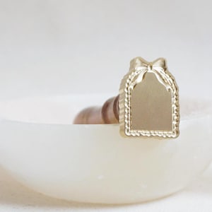 Puede incluir: Un sello dorado con un diseño rectangular y un lazo en la parte superior. El sello tiene un borde decorativo y está sobre un fondo blanco. El sello está sobre un cuenco blanco.