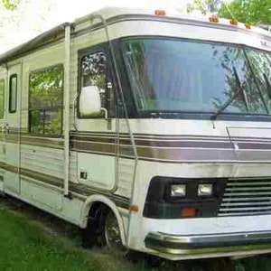 May include: A white and brown motorhome with a large windshield and a grill with a chrome bumper. The motorhome has a large awning and a side door.