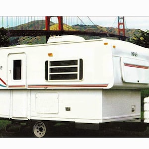 May include: A white TowLite camper with a red, white, and blue logo. The camper has a large window on the side and a smaller window on the back. The camper is parked in front of a green grassy area with a red bridge in the background.