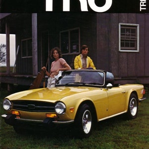 May include: A yellow Triumph TR6 convertible sports car parked in front of a wooden building. Two people are standing on the hood of the car.
