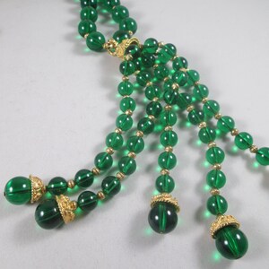 Pu&ograve; includere: Una lunga collana di perline di vetro verde smeraldo con dettagli dorati e un pendente a nappa. La collana &egrave; composta da pi&ugrave; file di perline e ha una chiusura dorata.