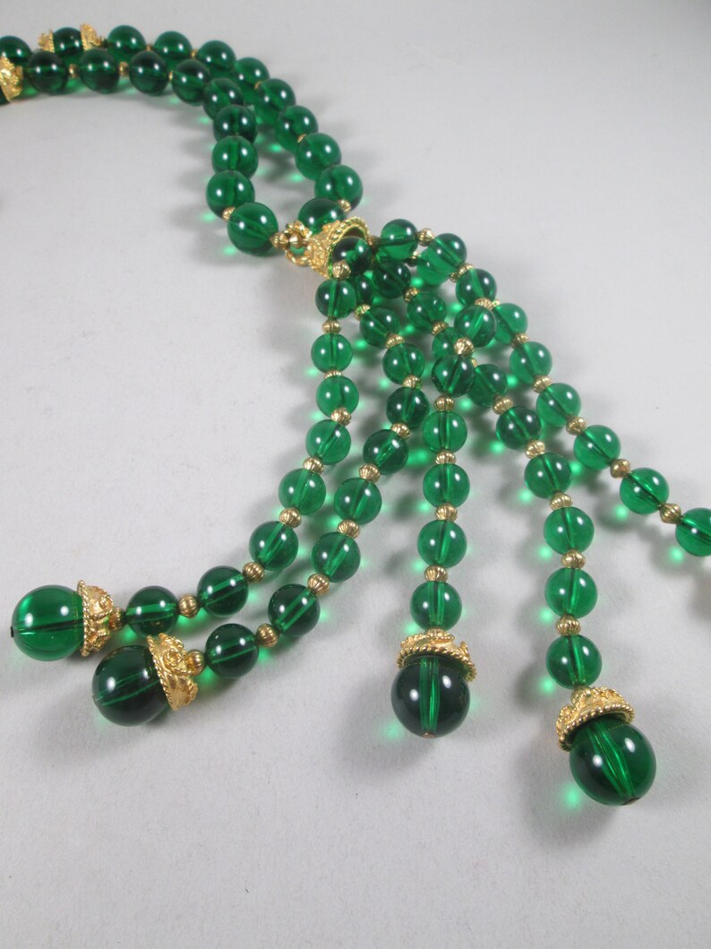 Pu&ograve; includere: Una lunga collana di perline di vetro verde smeraldo con dettagli dorati e un pendente a nappa. La collana &egrave; composta da pi&ugrave; file di perline e ha una chiusura dorata.