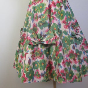 Op de afbeelding: Een witte jurk met een groene, roze en blauwe bloemenprint. De jurk heeft een strik op de rug en een geplooide rok.