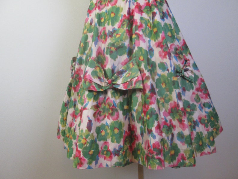 Op de afbeelding: Een witte jurk met een groene, roze en blauwe bloemenprint. De jurk heeft een strik op de rug en een geplooide rok.