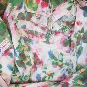 Op de afbeelding: Een witte stof met een roze, groene en blauwe bloemenprint. De stof heeft een rits en een label met de tekst "Creaciones Rafael ez Hecho en Mexico".