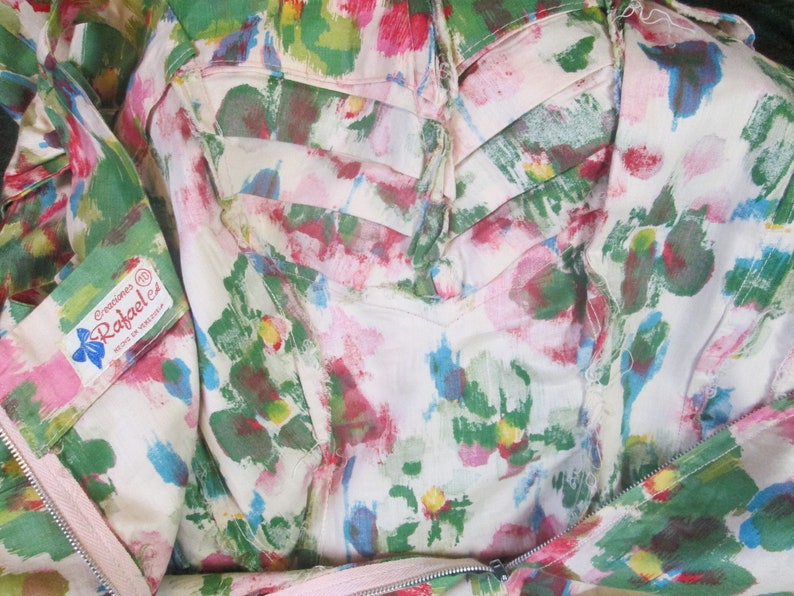 Op de afbeelding: Een witte stof met een roze, groene en blauwe bloemenprint. De stof heeft een rits en een label met de tekst "Creaciones Rafael ez Hecho en Mexico".