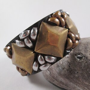 May include: A wide black cuff bracelet with a geometric design. The bracelet is decorated with gold and silver metal pieces in a braided pattern.