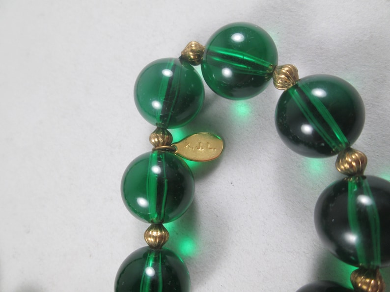 Pu&ograve; includere: Una collana di perline di vetro verde con accenti dorati. La collana ha un'etichetta che recita "KJL."