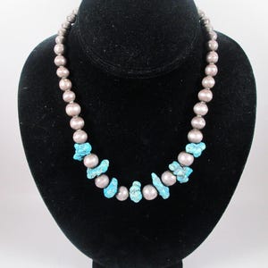 Puede incluir: Un collar con cuentas grises y detalles de piedra turquesa. El collar se exhibe sobre un busto de terciopelo negro. Las cuentas son redondas y espaciadas uniformemente, con piedras turquesas intercaladas.