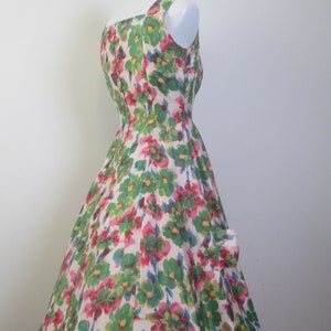 Op de afbeelding: Een vintage jurk zonder mouwen met een volle rok. De jurk is gemaakt van witte stof met een bloemenprint in roze, groene en blauwe tinten.