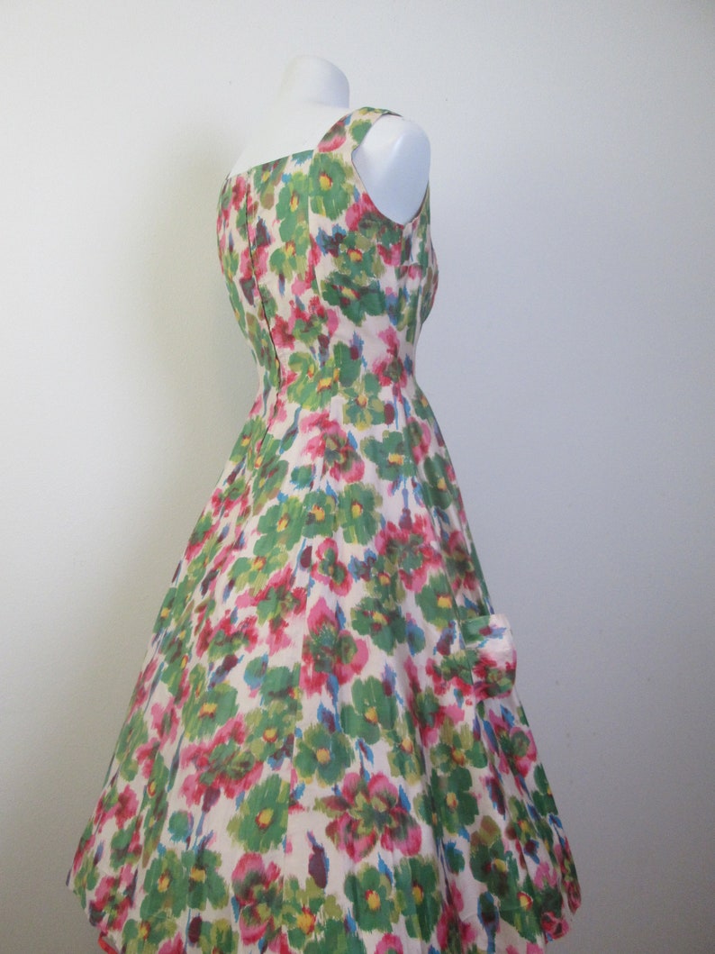 Op de afbeelding: Een vintage jurk zonder mouwen met een volle rok. De jurk is gemaakt van witte stof met een bloemenprint in roze, groene en blauwe tinten.