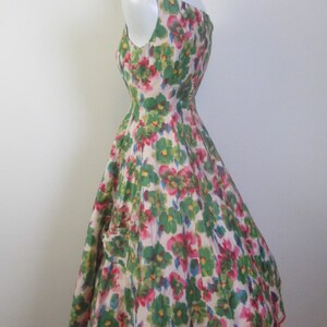 Op de afbeelding: Een mouwloze jurk met een witte achtergrond en een bloemenprint in tinten groen, roze en blauw. De jurk heeft een getailleerd bovenlijf en een volle rok.