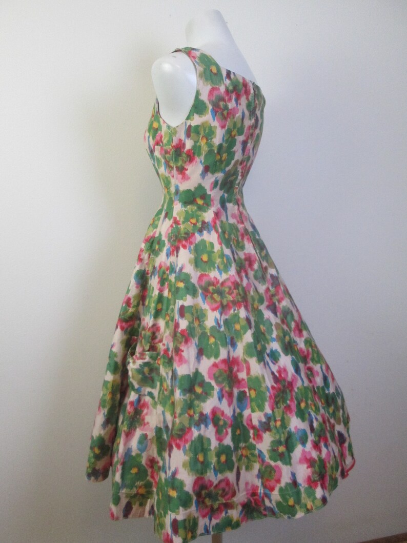 Op de afbeelding: Een mouwloze jurk met een witte achtergrond en een bloemenprint in tinten groen, roze en blauw. De jurk heeft een getailleerd bovenlijf en een volle rok.