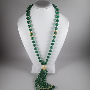 Pu&ograve; includere: Una lunga collana di perline di vetro verde smeraldo con un pendente dorato. La collana &egrave; su un espositore bianco.