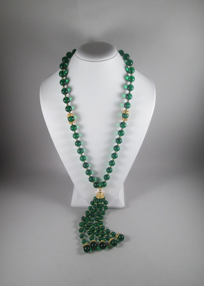 Pu&ograve; includere: Una lunga collana di perline di vetro verde smeraldo con un pendente dorato. La collana &egrave; su un espositore bianco.