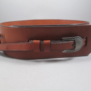 May include: A wide, brown leather belt with a silver-colored buckle. The belt has a decorative buckle and a matching silver-colored buckle on the belt strap. The belt is a western style.