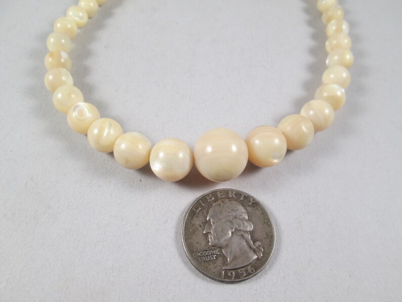 May include: A white pearl necklace with a diameter of approximately 1/2 inch. The necklace is laid out on a white surface with a US quarter for scale.