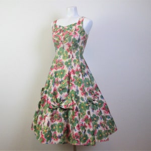 Op de afbeelding: Een vintage jurk met bloemenprint, een getailleerd bovenstuk en een volle rok. De jurk heeft een ronde hals en dunne bandjes. De stof is een lichtgewicht katoen met een bloemenpatroon in groen, roze en wit.