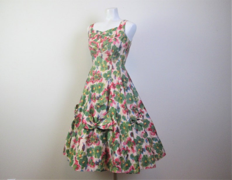 Op de afbeelding: Een vintage jurk met bloemenprint, een getailleerd bovenstuk en een volle rok. De jurk heeft een ronde hals en dunne bandjes. De stof is een lichtgewicht katoen met een bloemenpatroon in groen, roze en wit.