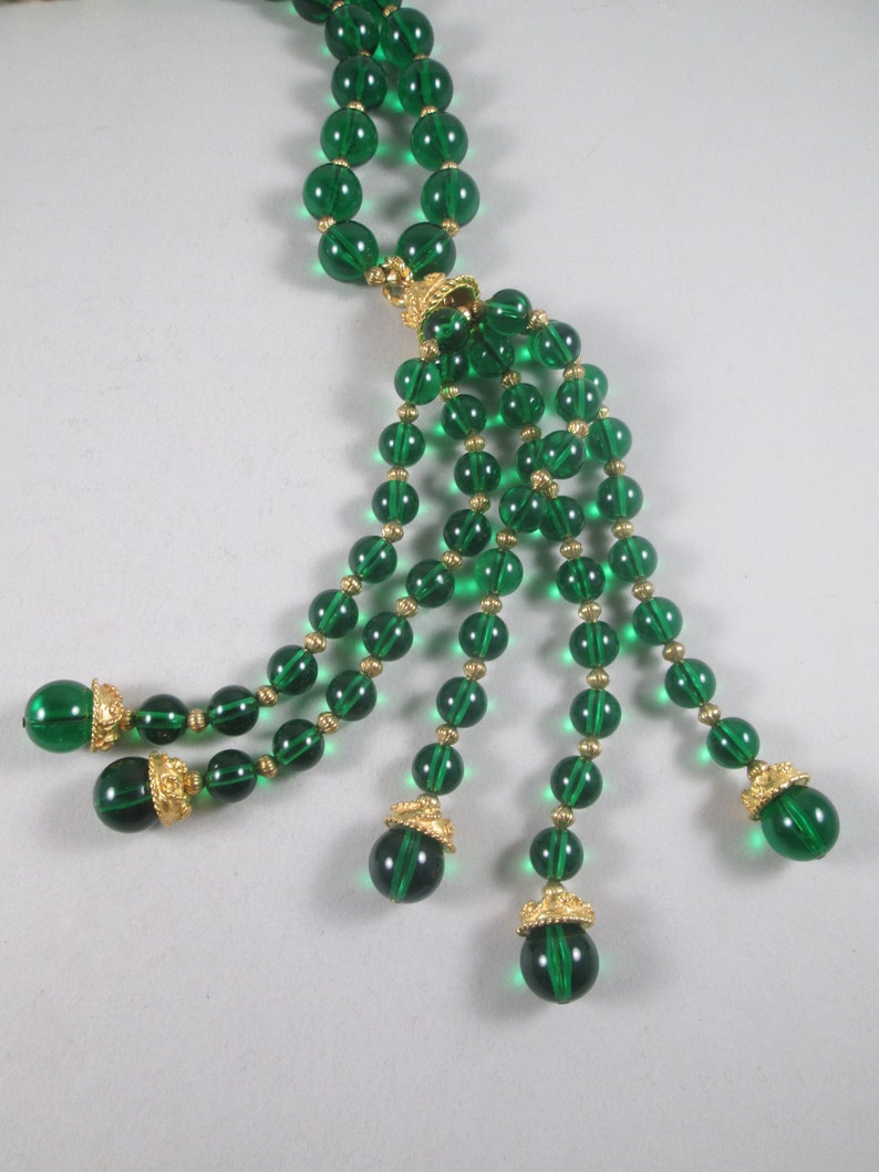 Pu&ograve; includere: Una lunga collana di perline di vetro verde smeraldo con un pendente di perline pi&ugrave; piccole. La collana ha una chiusura dorata e dettagli dorati sulle perline.