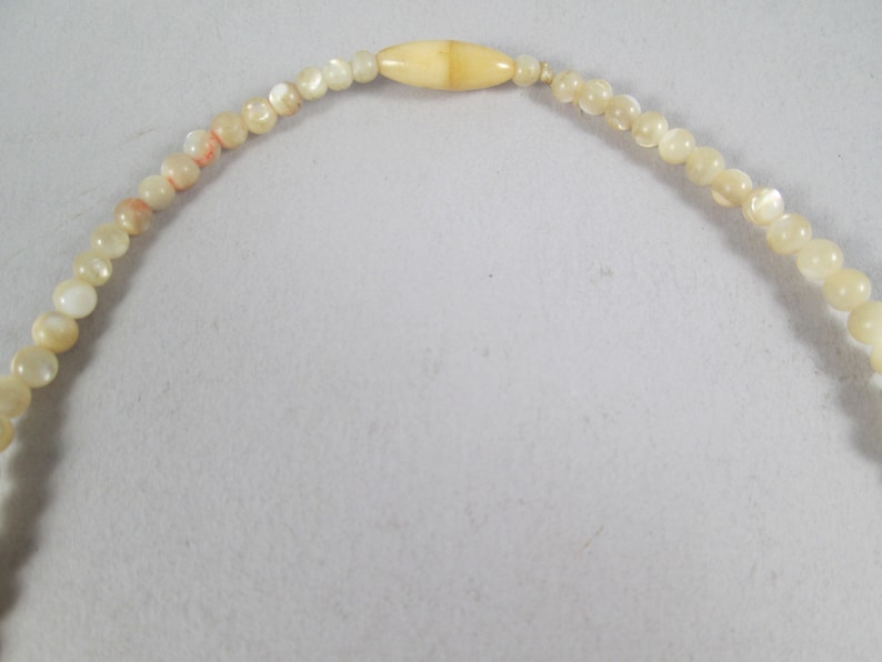 May include: A white and cream colored beaded necklace with a larger, oval-shaped bead in the center.