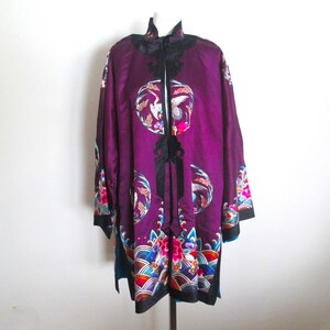 Puede incluir: Una chaqueta de manga larga en color morado intenso con cuello alto y ribetes negros. La prenda está decorada con motivos circulares bordados de grullas blancas y patrones florales coloridos, con bordados de ondas a lo largo del dobladillo.