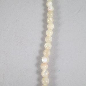 May include: A strand of white pearl beads with a subtle iridescent sheen. The beads have a delicate floral pattern and are strung on a clear thread.