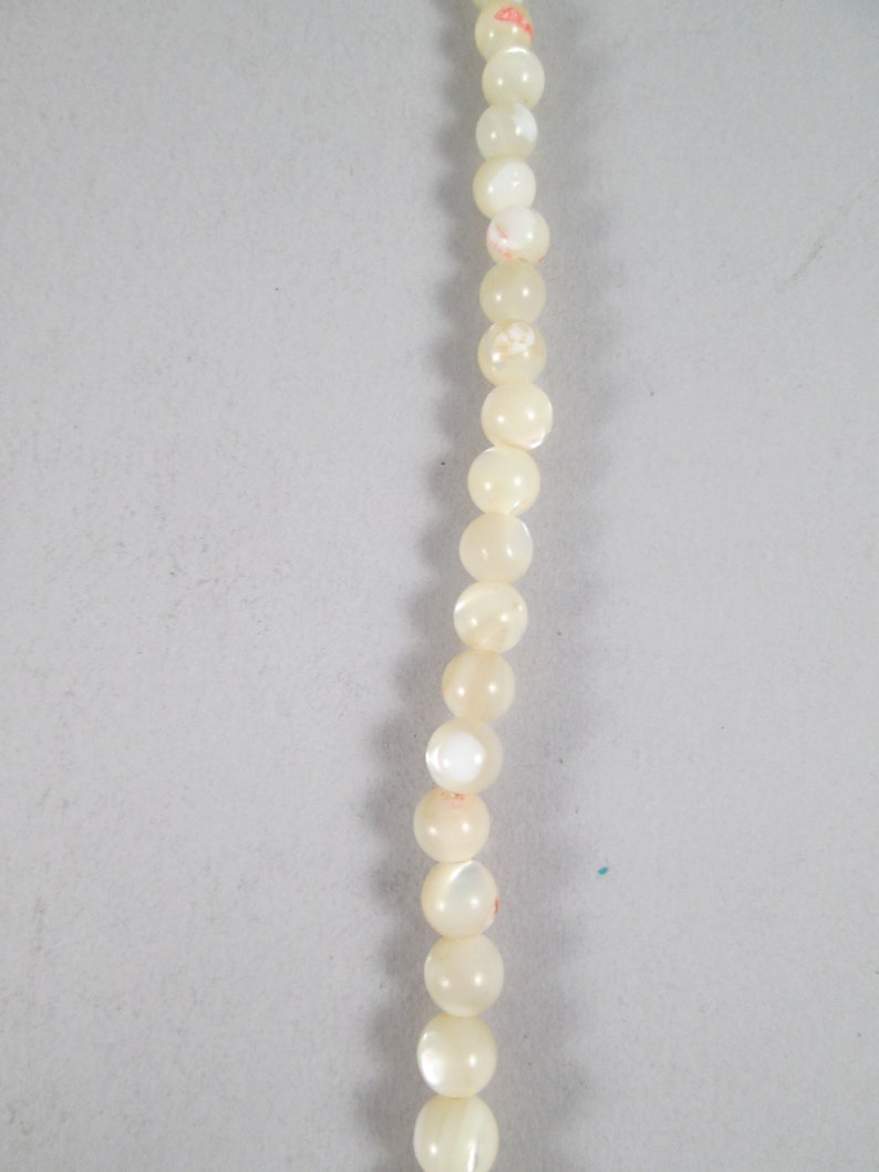 May include: A strand of white pearl beads with a subtle iridescent sheen. The beads have a delicate floral pattern and are strung on a clear thread.