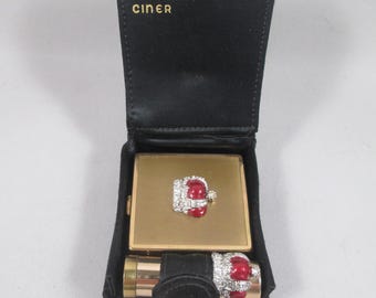 May include: A vintage CINER compact set in a black case. The set includes a gold-toned compact with a red and rhinestone embellishment, a lipstick holder, and a lipstick. The case is open, revealing the contents.