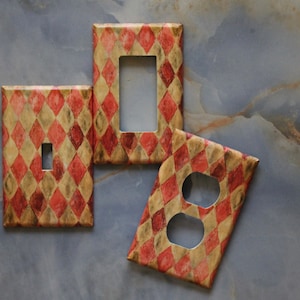 May include: Set of three decorative light switch covers. The covers feature a harlequin pattern in red and gold. Includes a single switch cover, a double switch cover, and an outlet cover.