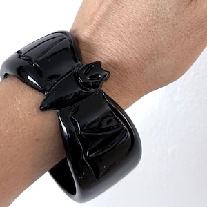 Puede incluir: Un brazalete negro brillante con un diseño de murciélago. El brazalete es ancho y rectangular, con un motivo de murciélago esculpido en el centro. El brazalete se lleva en una muñeca sobre un fondo blanco.