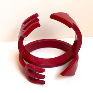 Puede incluir: Un par de brazaletes circulares de color rojo intenso con un diseño estilizado donde los extremos de cada banda tienen forma de tenedor y cuchara. El acabado liso y brillante resalta la estructura curva y abierta de estos accesorios.