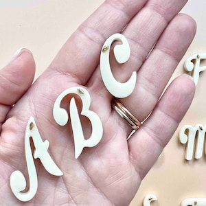 Op de afbeelding: Ivoor kleurige cursieve letterset, inclusief A, B en C, met kleine goudkleurige accenten. De letters worden weergegeven tegen een neutrale achtergrond, met andere letters zichtbaar op de achtergrond. De letters zijn waarschijnlijk voor knutsel- of decoratieve doeleinden.