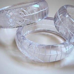 以下が含まれることがあります： クモの巣のデザインが施された、透明でクリアなバングル3つ。ブレスレットは透明な素材で作られており、クモと巣の模様が特徴です。バングルは明るく半透明な色をしています。