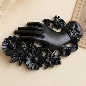 May include: A matte black brooch featuring a sculpted hand reaching out from a cluster of detailed flowers and leaves. The hand is adorned with a decorative cuff at the wrist, and the entire piece is finished in a solid, dark, opaque colour.