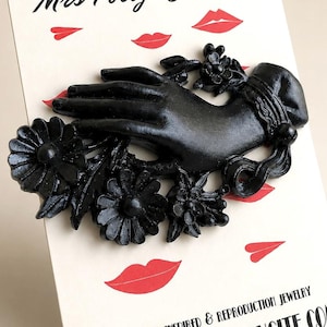 May include: A black brooch shaped like a hand adorned with flowers, mounted on a display card featuring the text Mrs Polly's Lucite, Vintage Inspired & Reproduction Jewelry, and www.mrspollyslucite.com with red lip illustrations.