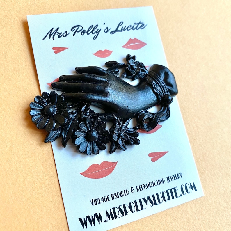 May include: A black brooch shaped like a hand adorned with flowers and a cuff, mounted on a white display card featuring the text Mrs Polly's Lucite, VINTAGE INSPIRED & REPRODUCTION JEWELRY, and WWW.MRSSPOLLYSLUCITE.COM with small pink lip icons.