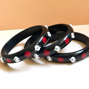 Op de afbeelding: Drie zwarte armbanden met een herhalend patroon van kleine witte spookfiguren en effen rode cirkelvormige stippen. De armbanden hebben een gladde, glanzende afwerking en een afgerond profiel, met een consistent ontwerp op alle drie de stukken.