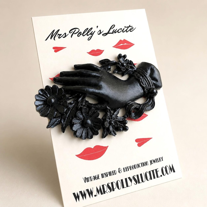 May include: A black brooch shaped like a hand adorned with flowers, mounted on a display card featuring the text Mrs Polly's Lucite, Vintage Inspired & Reproduction Jewelry, and www.mrspollyslucite.com with red lip illustrations.