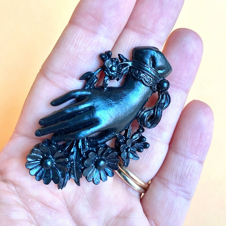 May include: A matte black brooch shaped like a hand adorned with a decorative cuff and surrounded by sculpted floral elements. The hand is positioned with fingers extended, featuring intricate detailing on the flowers and the wrist cuff design.