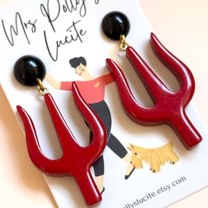 May include: A pair of dangle earrings featuring glossy red trident-shaped charms suspended from black circular studs. The earrings are displayed on a white card printed with the text Mrs. Polly's Lucite and mrspollyslucite.etsy.com, along with an illustration of a person and a dog.