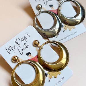 May include: Two pairs of hoop earrings, one silver and one gold, displayed on white cards. The earrings have a large, rounded design. The cards feature the text "Mrs Polly's Lucite" and a website address.