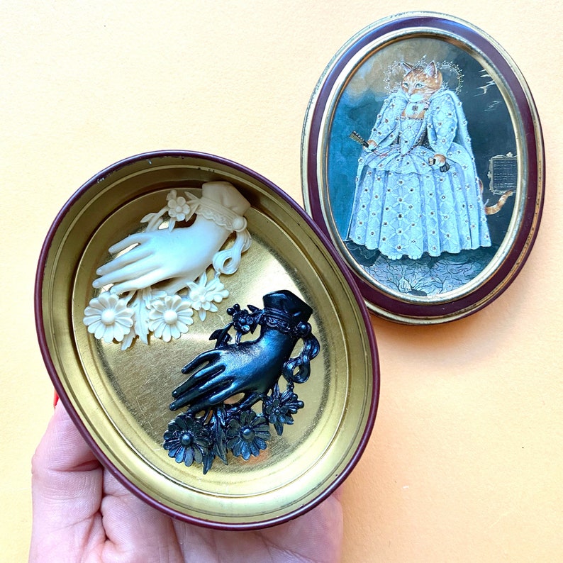 May include: An oval metal tin with a maroon rim and a lid featuring a cat dressed in a royal gown. Inside the tin are two hand-shaped brooches, one in white and one in black, each adorned with floral details and lace-like cuffs at the wrist.