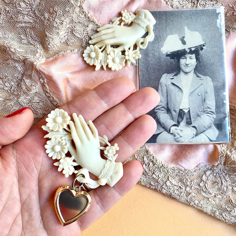 May include: A cream-coloured brooch featuring a sculpted hand adorned with flowers and a dangling gold-tone heart-shaped locket. The brooch is paired with a black and white vintage photograph of a person wearing a hat, set against a lace fabric background.