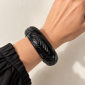 May include: A wide, glossy black bangle bracelet featuring a carved, repeating leaf pattern that wraps around the entire circumference of the piece. The surface is smooth and reflective, showcasing intricate, recessed detailing.