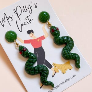 May include: A pair of dark green snake-shaped earrings with red crystal eyes and a textured scale pattern. The earrings are attached to a white display card featuring the text Mrs Polly's Lucite and mrspollyslucite.etsy.com with an illustration.