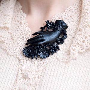 May include: A matte black brooch shaped like a hand emerging from a ruffled cuff, surrounded by detailed floral accents including several daisy-like flowers and small buds. The piece features a smooth, dark finish and intricate sculptural details.