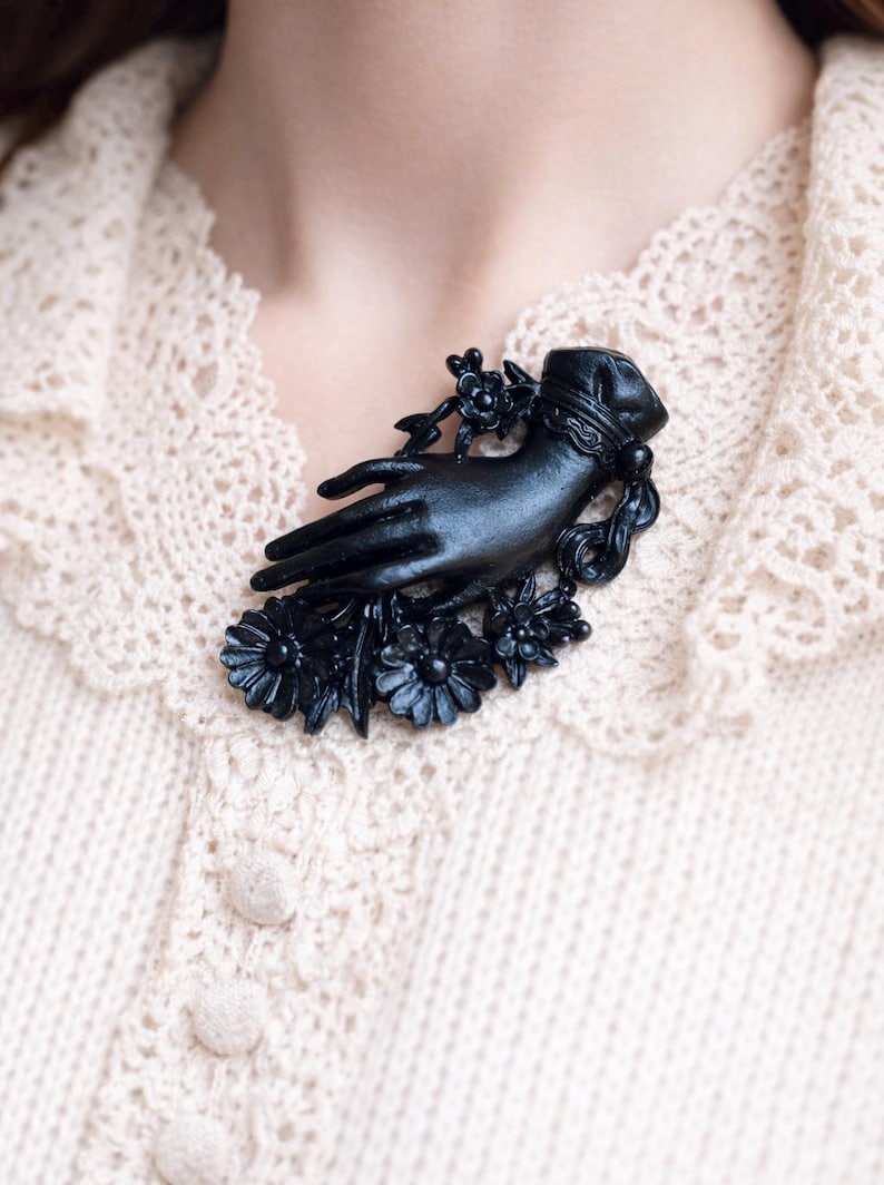May include: A matte black brooch shaped like a hand emerging from a ruffled cuff, surrounded by detailed floral accents including several daisy-like flowers and small buds. The piece features a smooth, dark finish and intricate sculptural details.